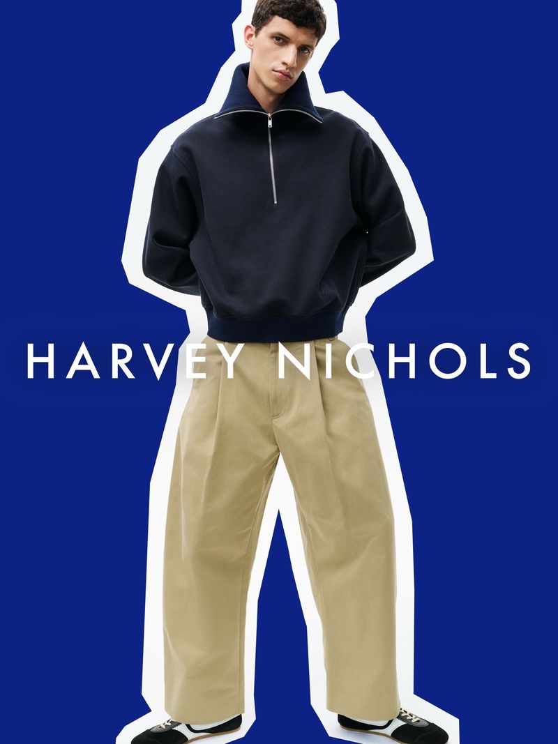 Model wearing a dark half-zip sweatshirt and pleated khaki trousers against a vibrant blue cutout background.