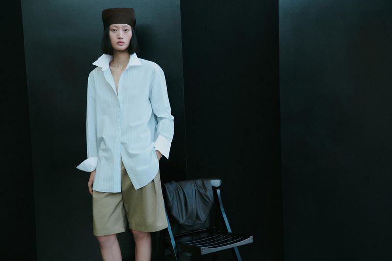 Model Yilan Hua wearing an oversized collared shirt, khaki shorts, and a brown headwrap next to a chair against a black background.