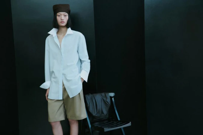 Model Yilan Hua wearing an oversized collared shirt, khaki shorts, and a brown headwrap next to a chair against a black background.