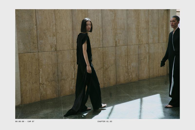 Two female models in black outfits walking in a hallway with stone tile walls and natural light casting bright angular shadows.
