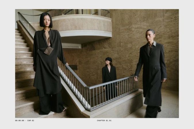 Three female models in dark attire standing on stone stairs inside a grand building with marble walls and railings.
