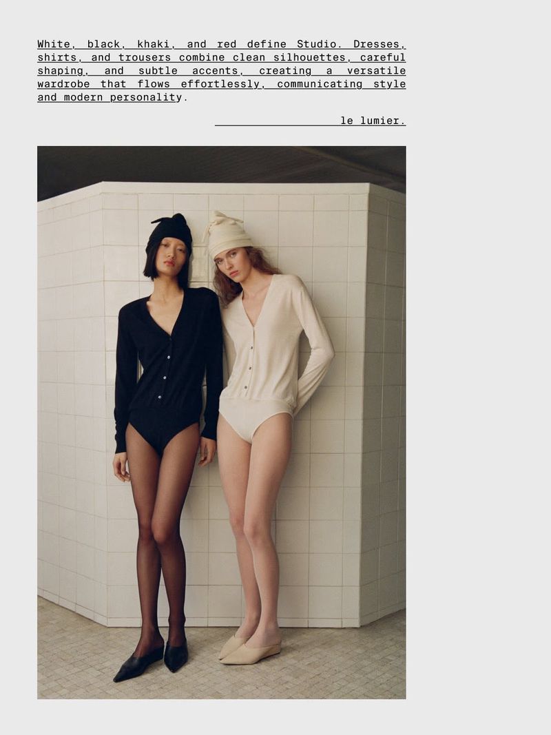 Two models in black and cream knit bodysuits and matching beanies standing against a tiled partition wall.