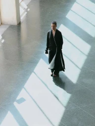 High angle of a woman walking across a grey tiled floor bathed in geometric sunlight, wearing a long black coat with white lining.
