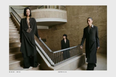 Three female models in dark attire standing on stone stairs inside a grand building with marble walls and railings.