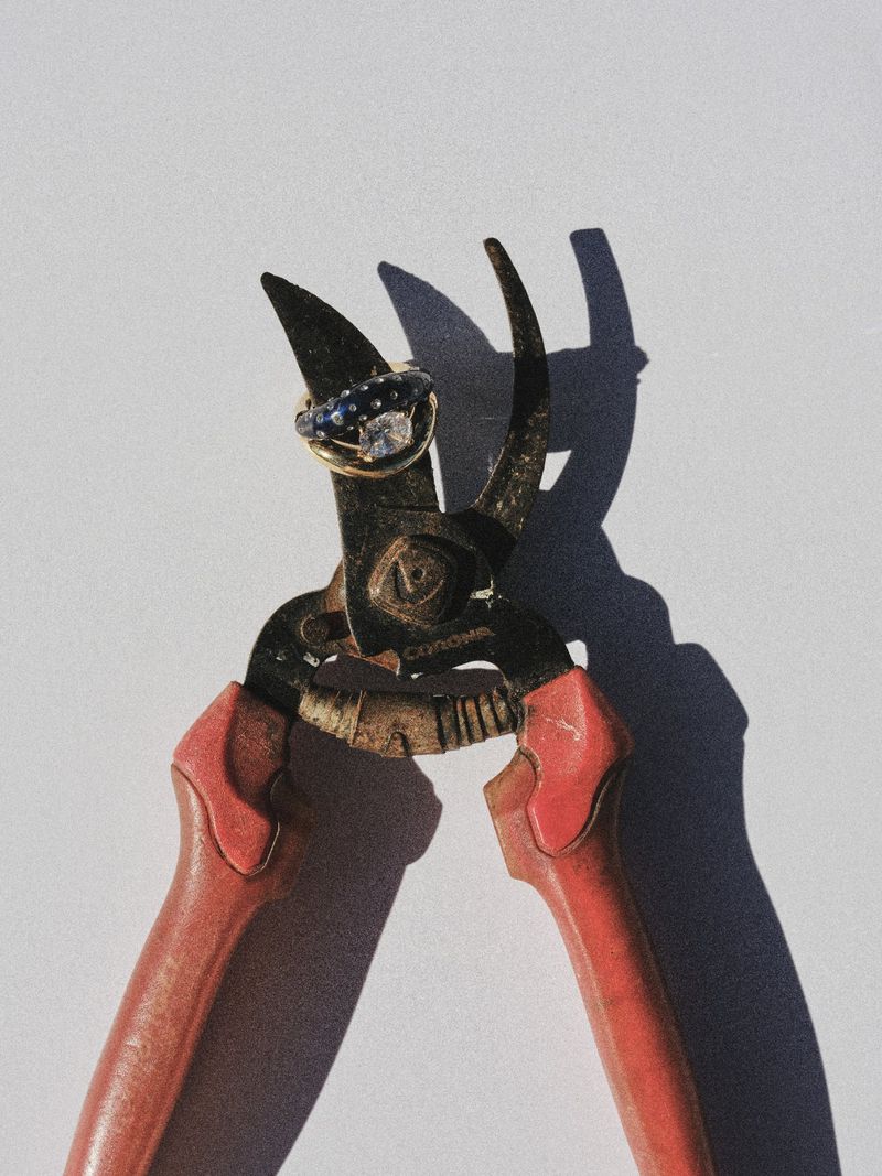 Red-handled pruning shears holding a diamond and sapphire ring against a white surface in harsh direct sunlight.