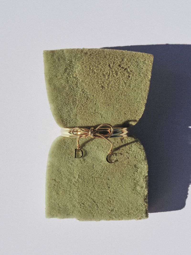 Gold bow ring with D and C letter charms cinching a textured olive green sponge against a white backdrop with a hard shadow.