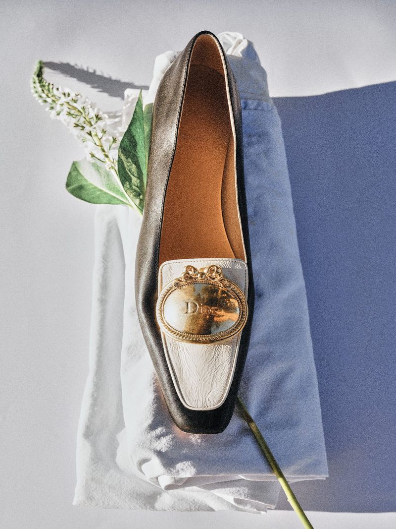 Two-tone black and white leather Dior loafer with a gold emblem resting on folded white fabric beside a white flower stem.
