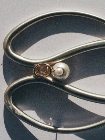 Close-up of a gold logo charm and pearl earring placed on a curved matte silver metallic sculpture against a pale backdrop.