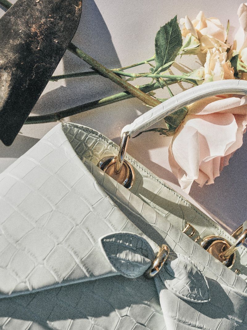 Close-up of a pale mint crocodile-embossed leather handbag arranged with pale pink roses and a garden trowel under direct sunlight.