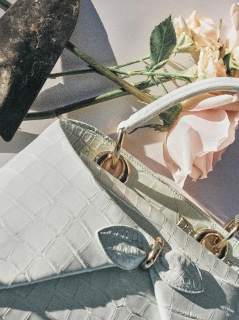 Close-up of a pale mint crocodile-embossed leather handbag arranged with pale pink roses and a garden trowel under direct sunlight.