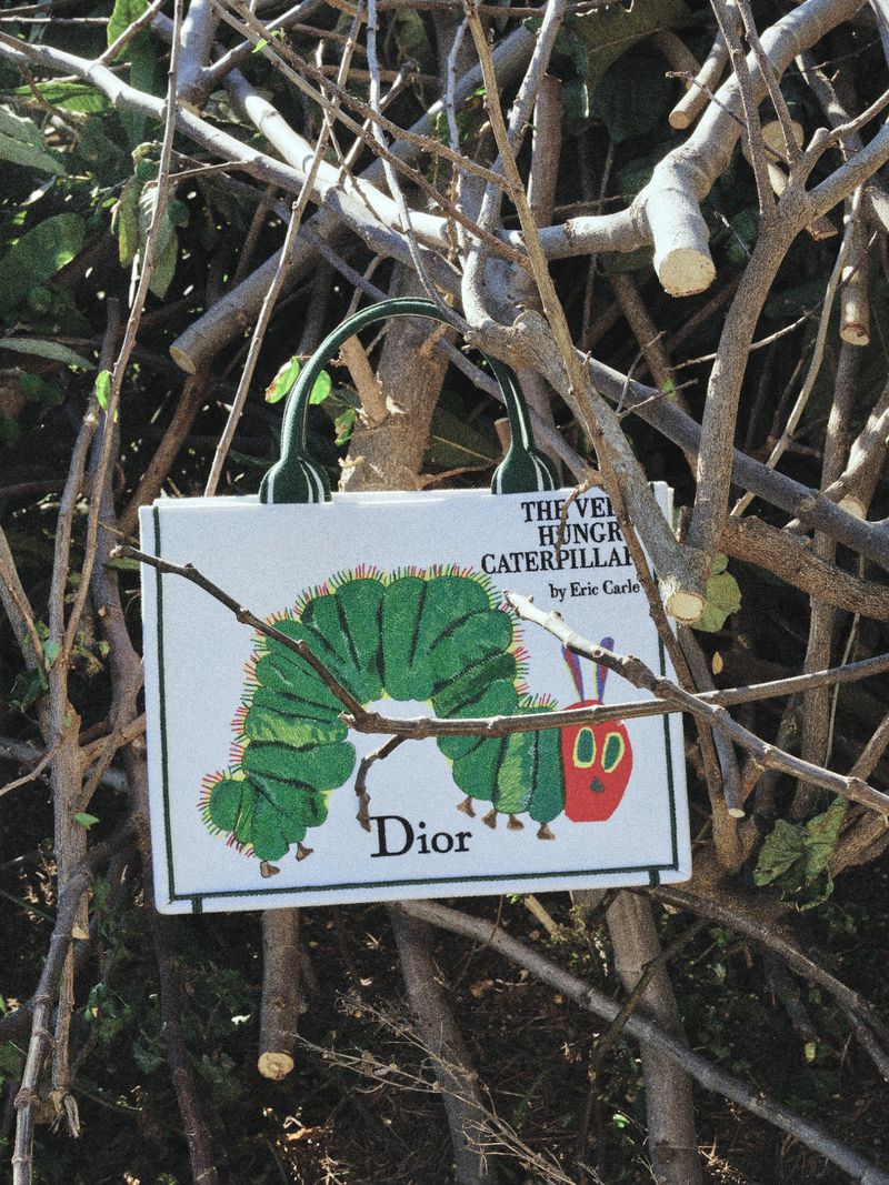 Dior tote bag featuring The Very Hungry Caterpillar illustration hanging in tangled tree branches outdoors in direct sunlight.