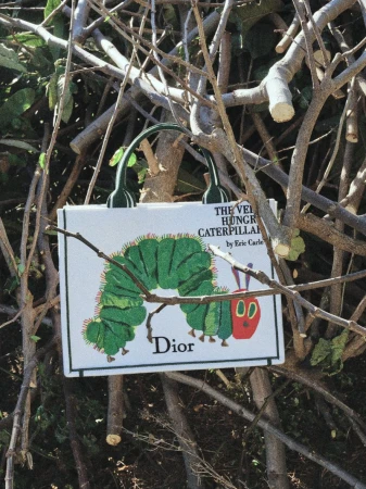 Dior tote bag featuring The Very Hungry Caterpillar illustration hanging in tangled tree branches outdoors in direct sunlight.