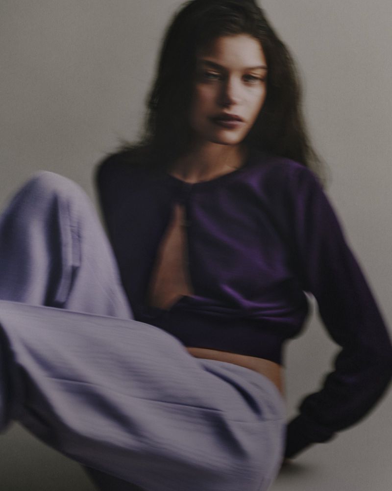 Model wearing a deep purple top and light purple trousers posed against a plain grey background