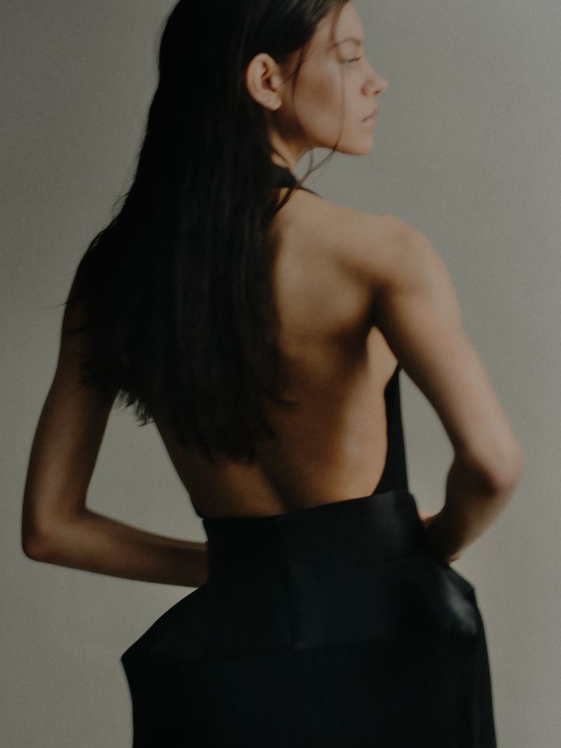 Model facing away in a black backless dress with a wide structured waist against a neutral studio backdrop.