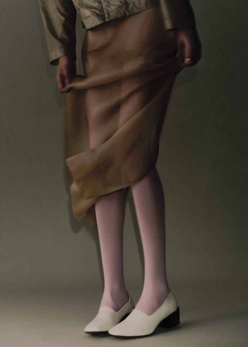 Lower-body view of a model lifting a sheer brown skirt, paired with white loafers and pale tights against a neutral studio backdrop.