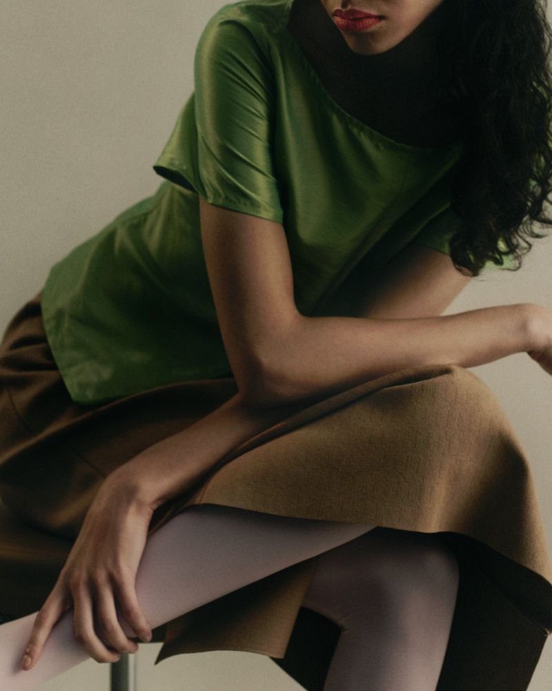Close-up of a woman seated wearing a green satin short-sleeved top, camel brown skirt, and pale tights in front of a studio backdrop.