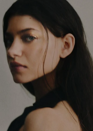 Close-up portrait of a woman with dark hair looking over her bare shoulder against a neutral background.