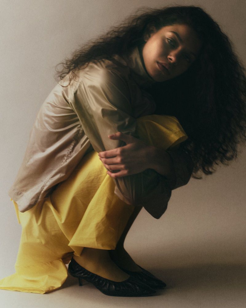 Model with long dark curls crouching in a yellow skirt, taupe high-neck top, and black kitten heels against a neutral studio backdrop.