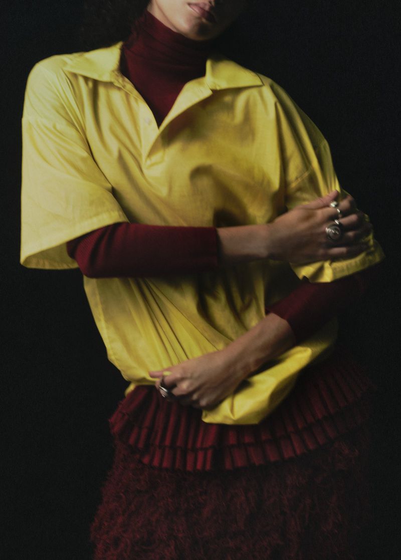 Model wearing an oversized yellow short-sleeve polo shirt layered over a burgundy turtleneck against a black background.