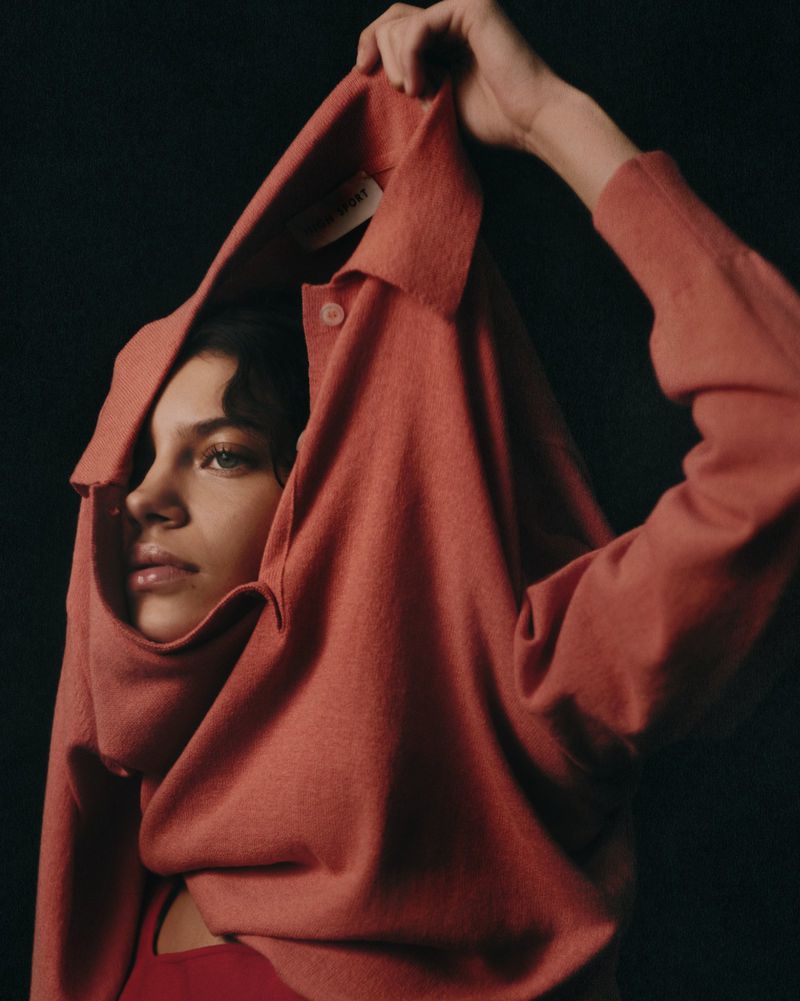 Young woman in studio portrait, wearing casual clothes and holding a reddish-brown collared shirt over her head, looking contemplative.