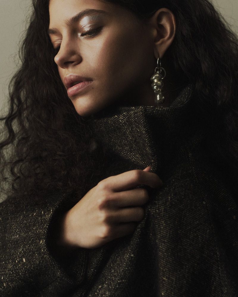 Close-up of a woman in a dark high-collar tweed coat wearing a sculptural silver drop earring against a neutral backdrop.