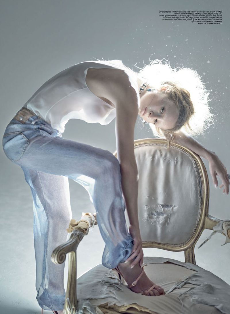 Model Maria Klaumann poses arched over a distressed antique armchair wearing sheer trousers and a white top against a studio background.