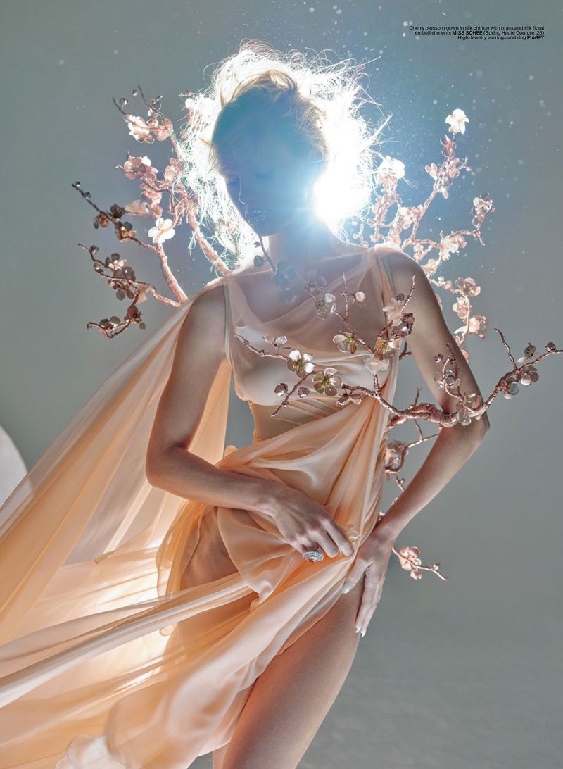 Model Maria Klaumann poses in a draped peach chiffon gown with sculptural flowering branches under a radiant halo backlight.