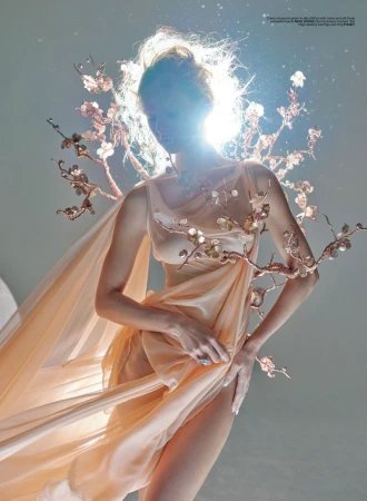 Model Maria Klaumann poses in a draped peach chiffon gown with sculptural flowering branches under a radiant halo backlight.