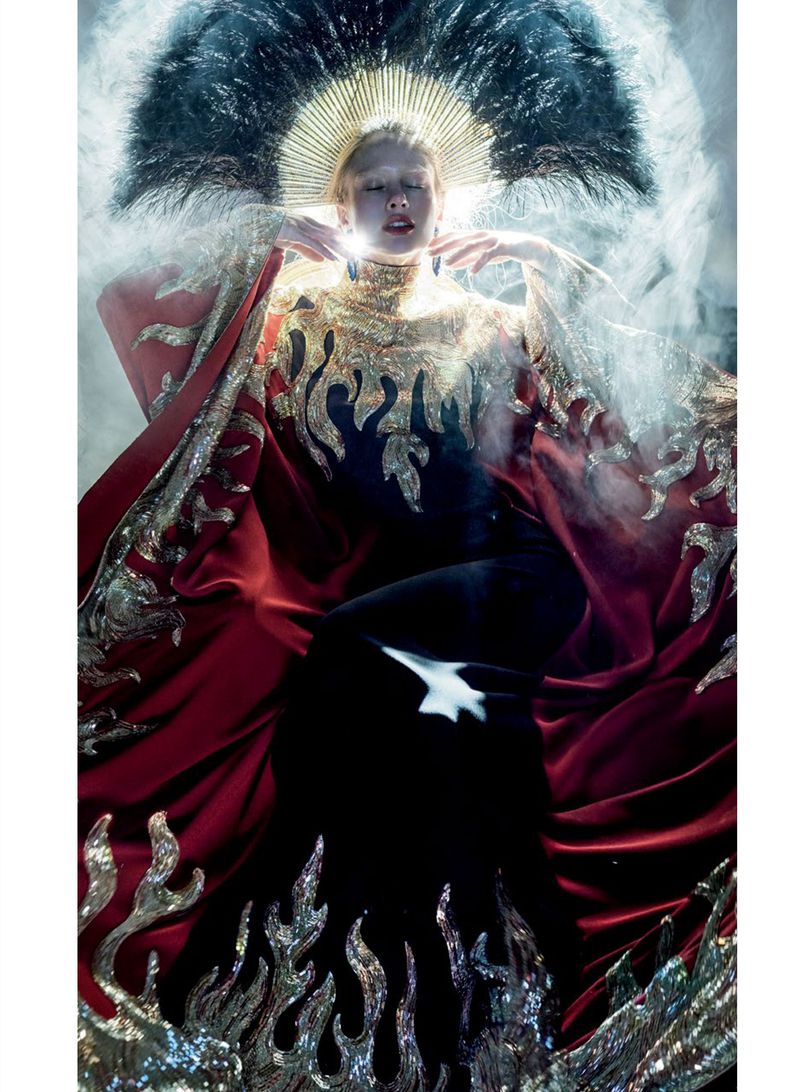 Model Maria Klaumann wearing an elaborate gold-embroidered crimson cape and a feather halo headdress amid ambient mist.