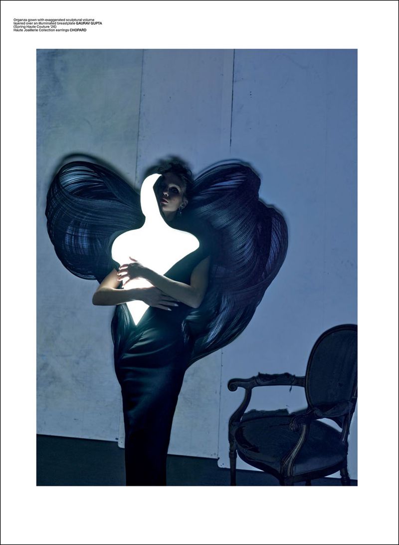 Maria Klaumann wearing a sculptural black gown with large wing-like accents while holding a glowing light form next to an antique chair.