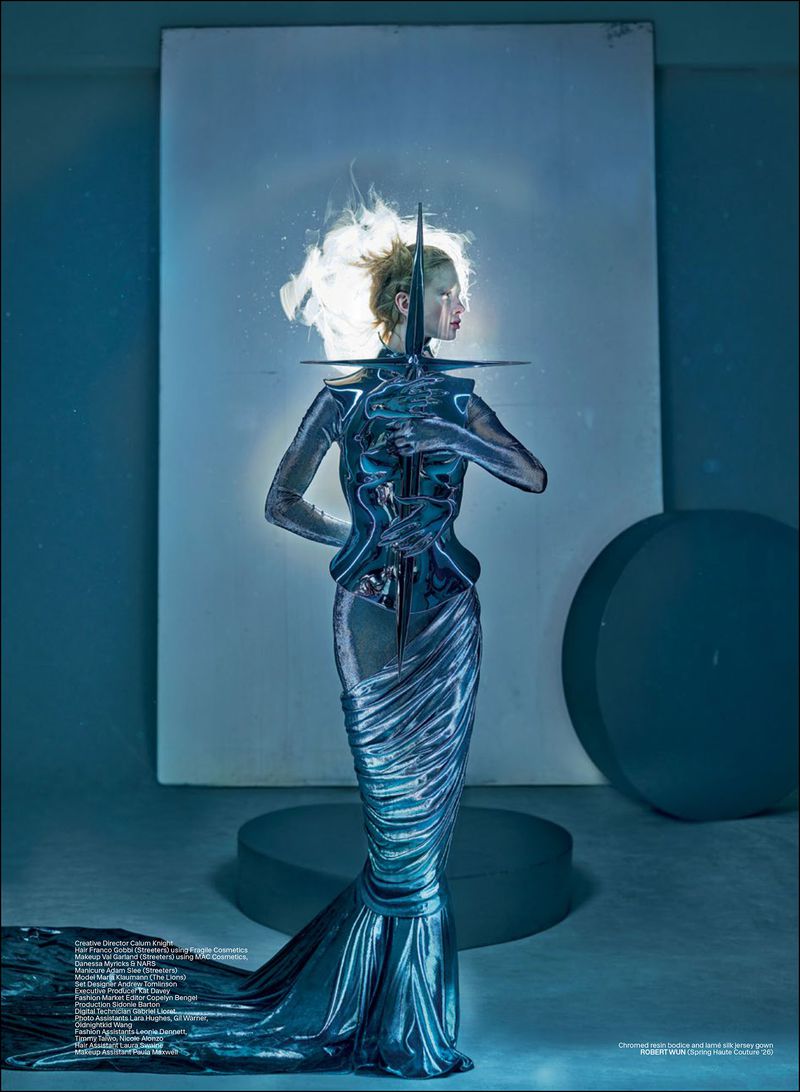 Model Maria Klaumann poses in a sculpted chrome corset and metallic silver fishtail gown in a cool-toned studio setting.