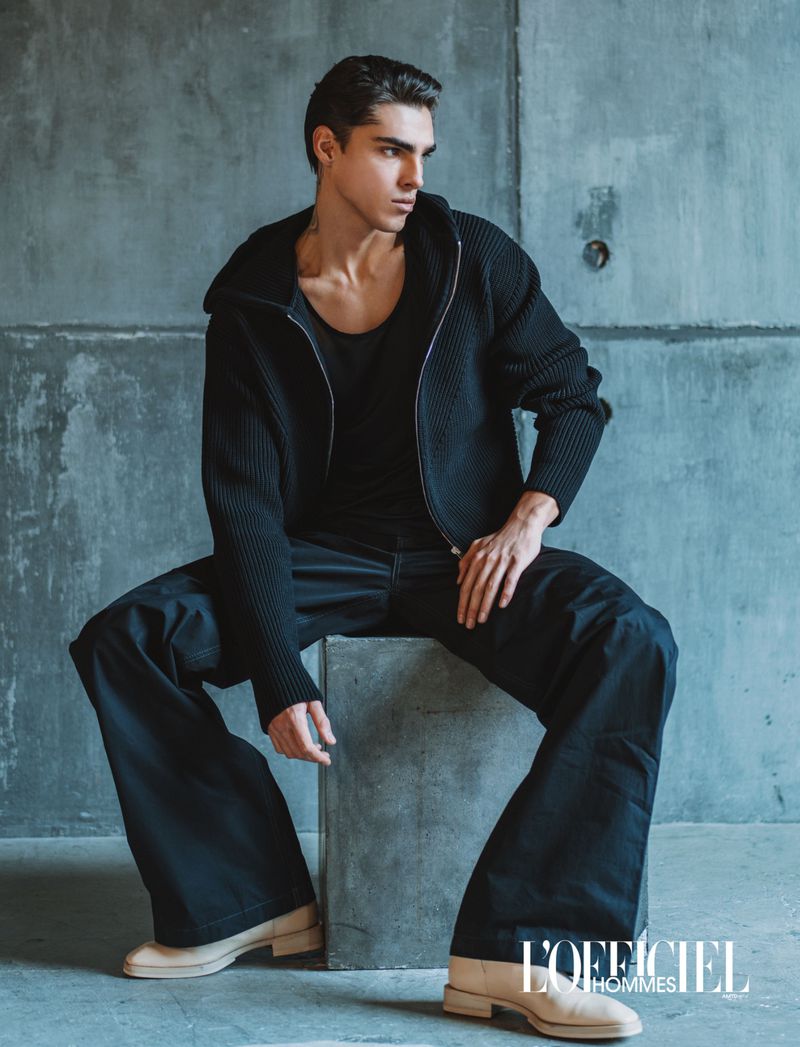 Model Simone Stravolo seated on a concrete cube wearing a black knit zip jacket, wide-leg trousers, and light beige boots.