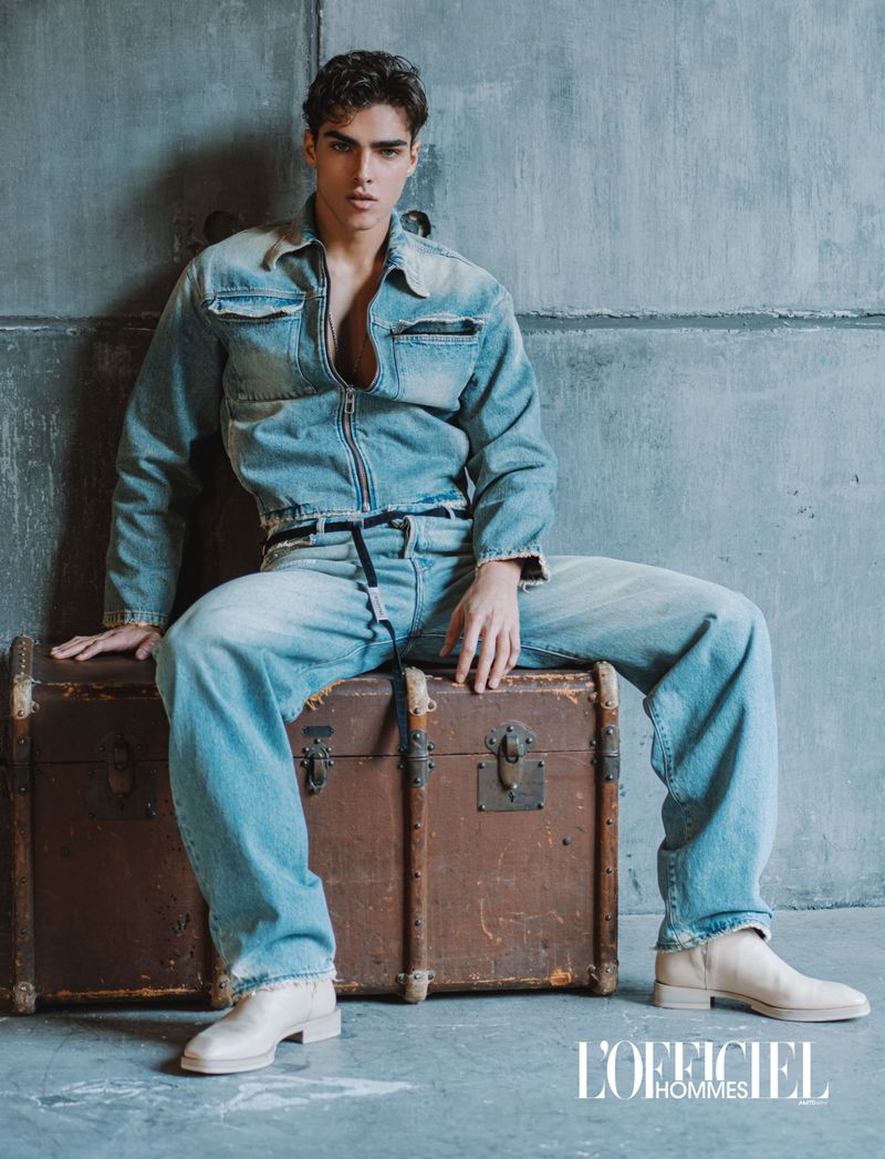 Simone Stravolo in a double denim outfit seated on a vintage brown trunk against a concrete wall.