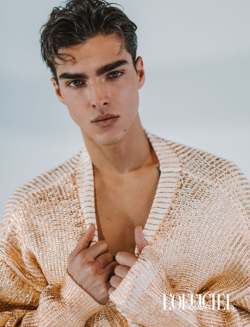 Model Simone Stravolo wearing an open metallic gold knit cardigan against a bright studio background for L'Officiel.
