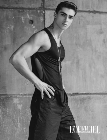 Model Simone Stravolo posing in a black sleeveless tank top and dark trousers against a concrete wall for L'Officiel Hommes.