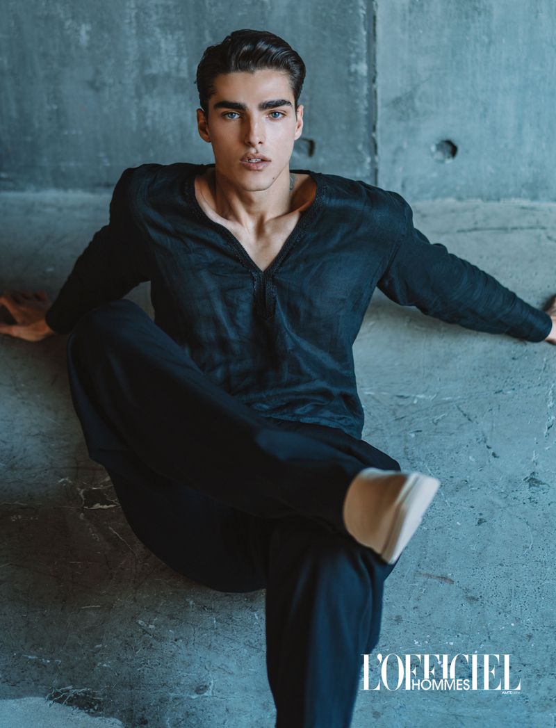 Male model Simone Stravolo seated on a concrete floor wearing a black top and dark trousers for L'Officiel Ukraine editorial.