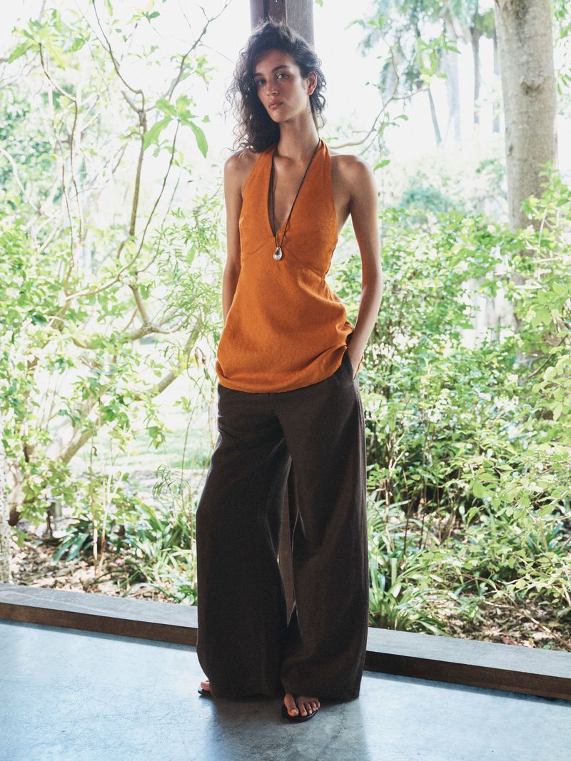 Model Lily Vander Meeden posing in an orange halter top and brown wide-leg pants against a lush garden backdrop for Aritzia.