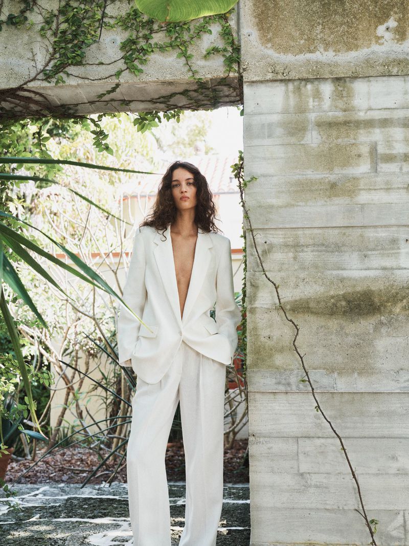 Model in a white linen pantsuit standing beneath a concrete architectural archway surrounded by lush greenery.