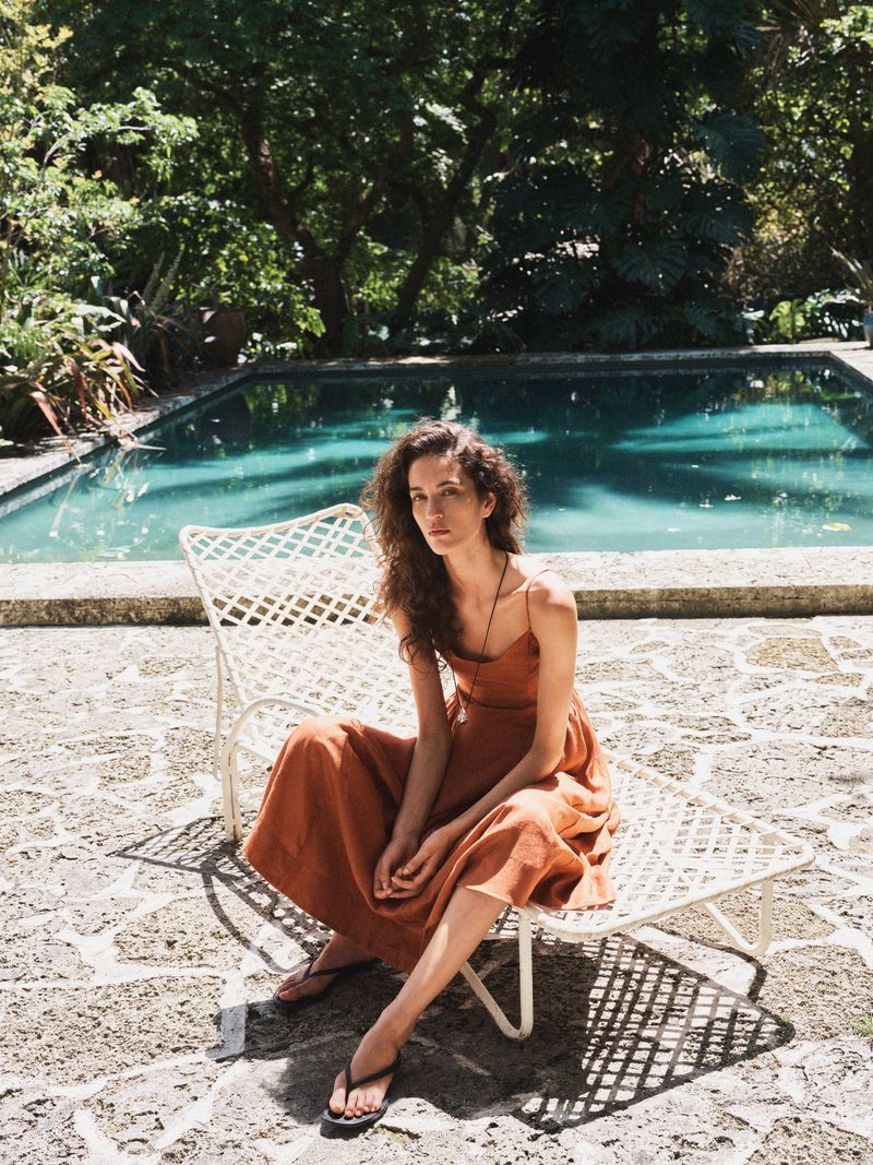 Lily Vander Meeden seated on a white lattice lounge chair wearing a rust orange linen dress beside an outdoor swimming pool.