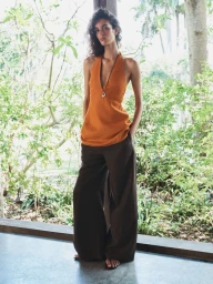 Model Lily Vander Meeden posing in an orange halter top and brown wide-leg pants against a lush garden backdrop for Aritzia.