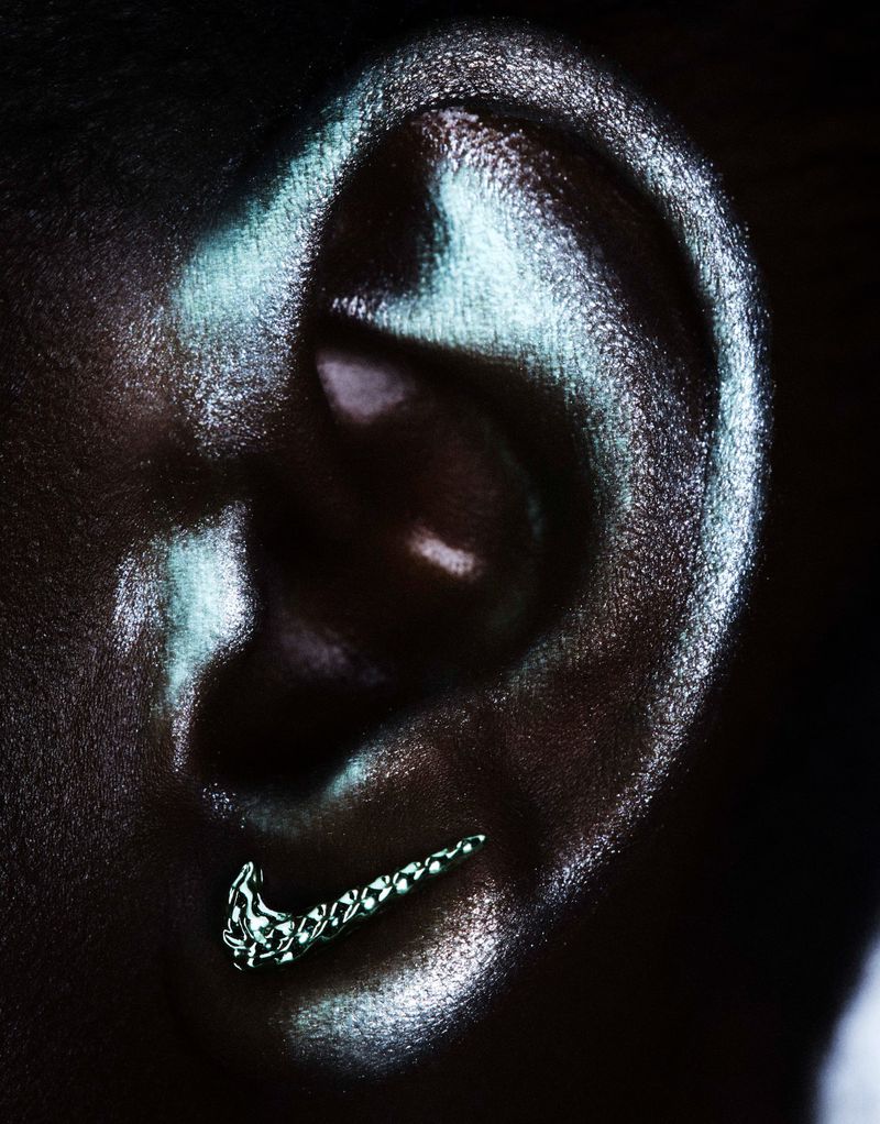 Close-up of a human ear covered in shimmering metallic makeup, wearing a textured silver swoosh-shaped earring on the lobe.