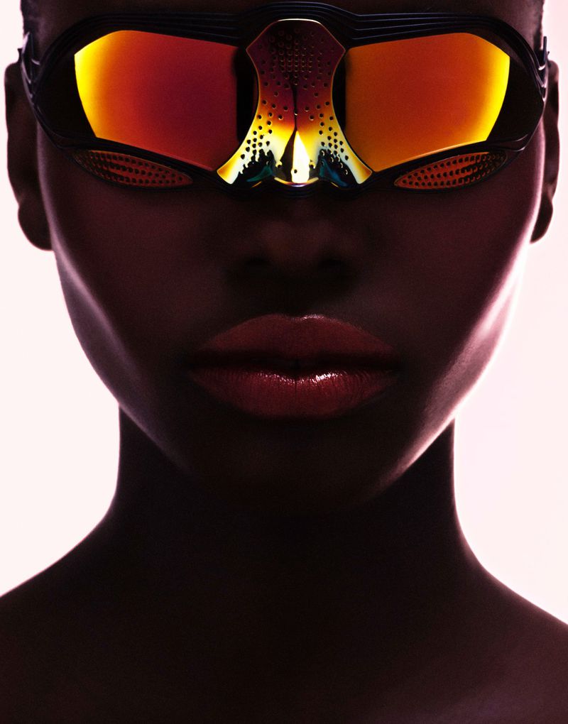 Close-up portrait of a Black woman wearing futuristic mirrored sunglasses with amber-orange lenses and glossy lips.