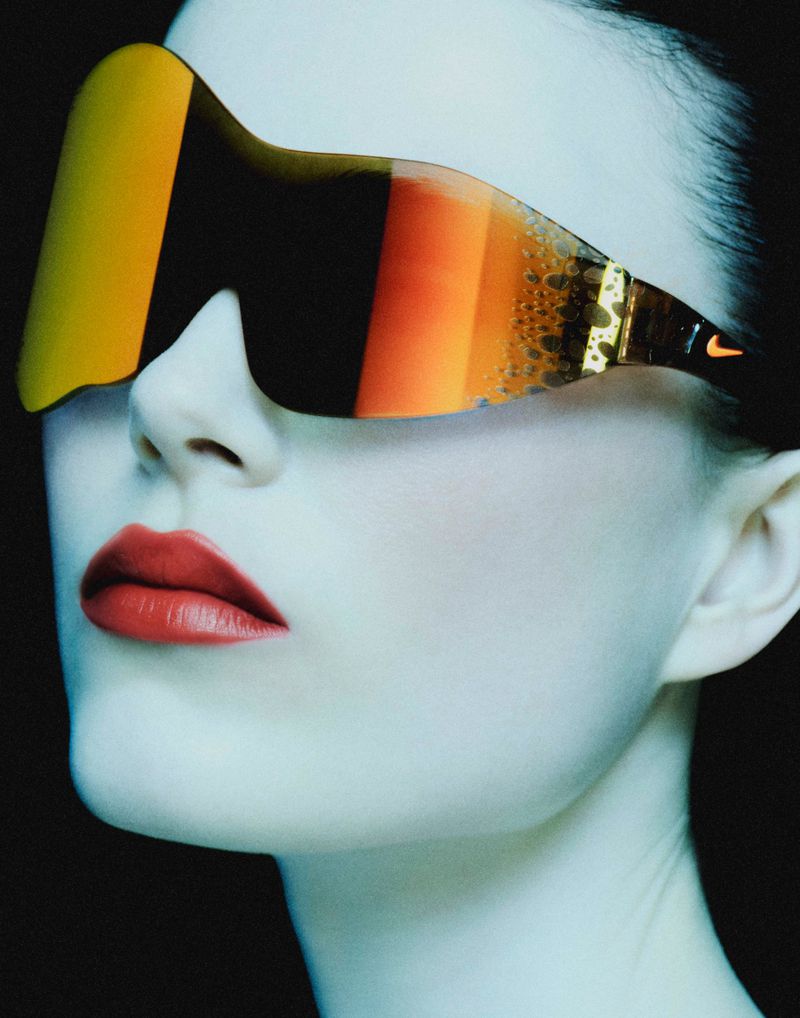 Close-up of a model wearing oversized orange shield visor sunglasses and bright red lipstick against a dark backdrop.