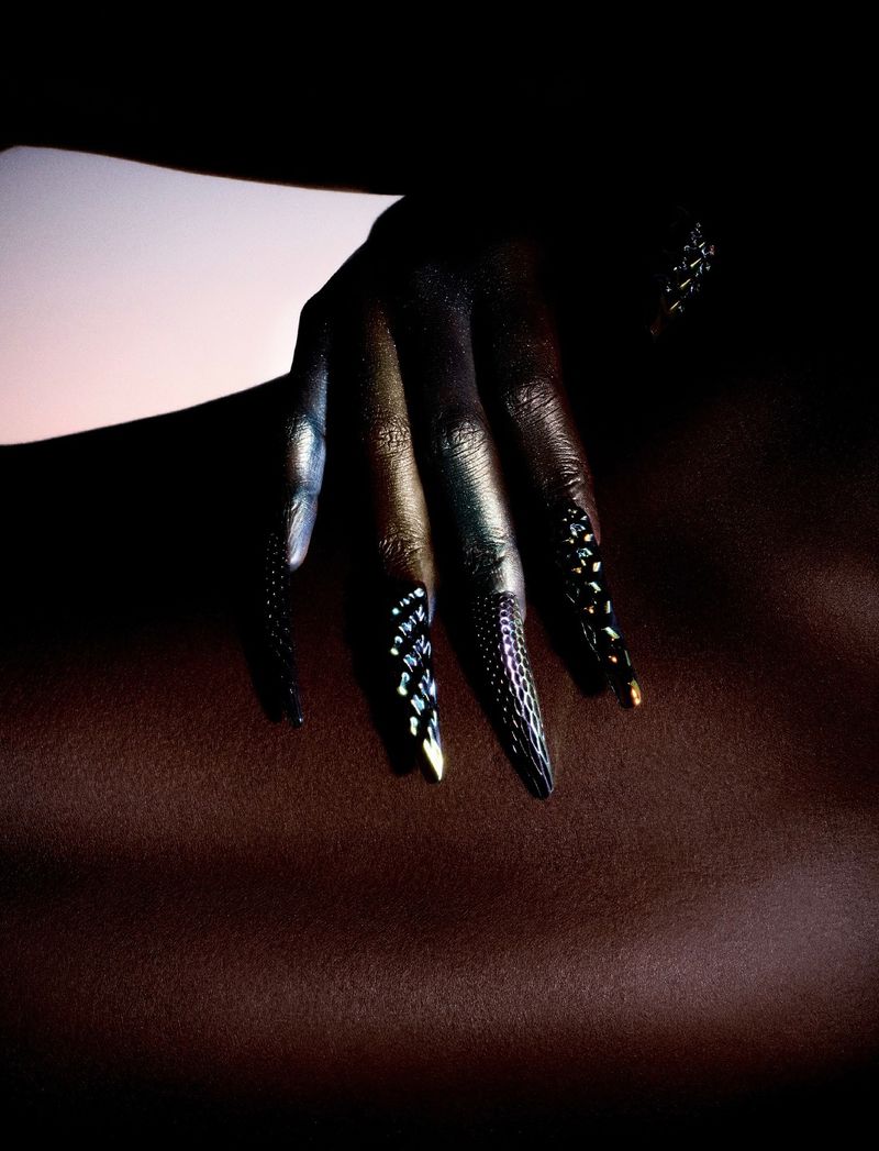 Close-up of a hand with iridescent metallic pigment and long, textured black stiletto nails resting on dark skin.