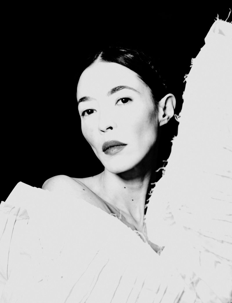 Close-up portrait of a woman with dark hair wearing structured white clothing against a black background.