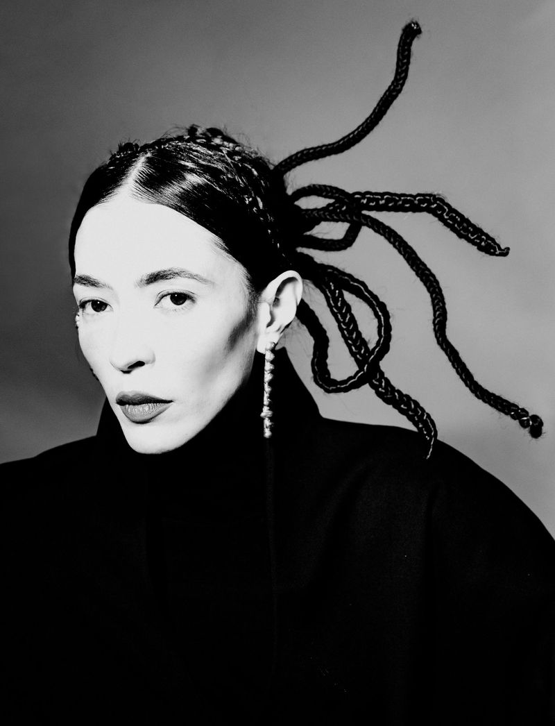 Black and white portrait of a woman with sculptural extended braids and a dangling earring wearing a high-neck black coat.