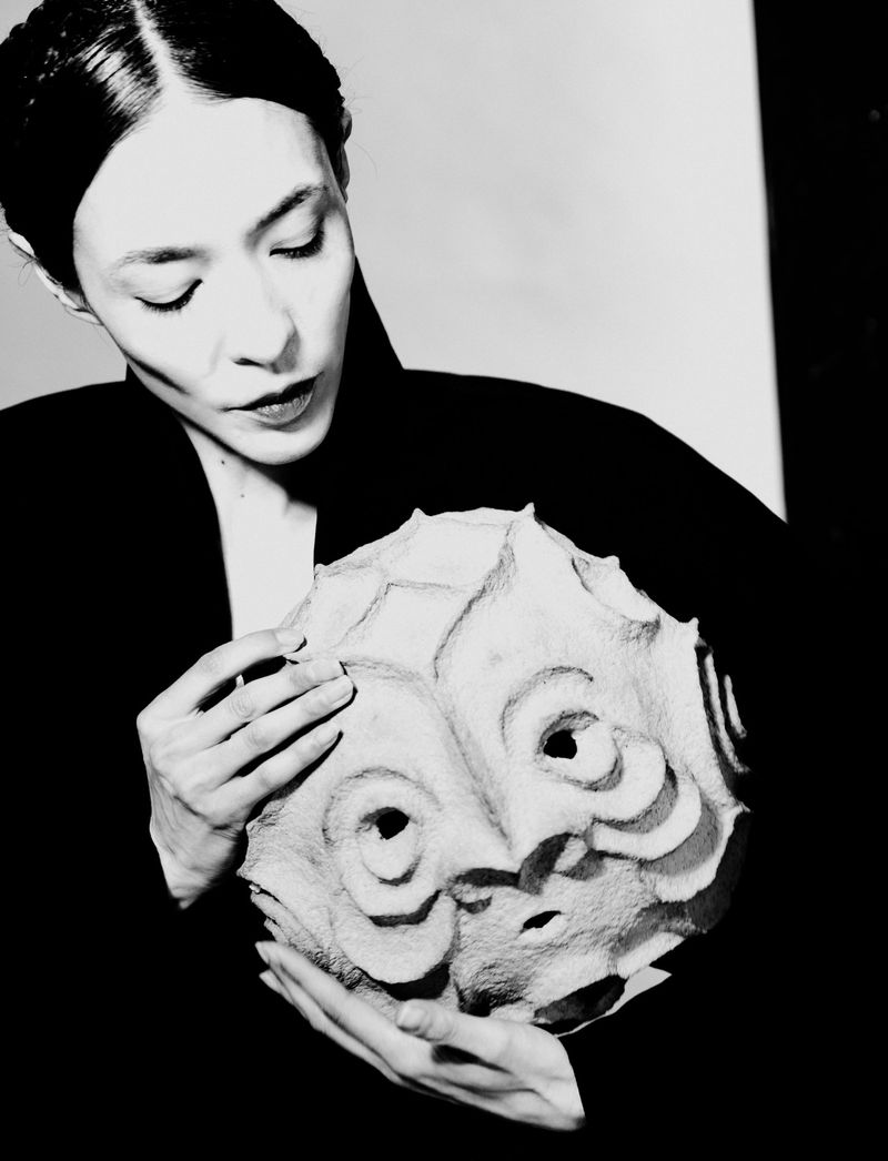 Woman looking down while holding a textured spherical sculpted mask in front of a high-contrast studio background.