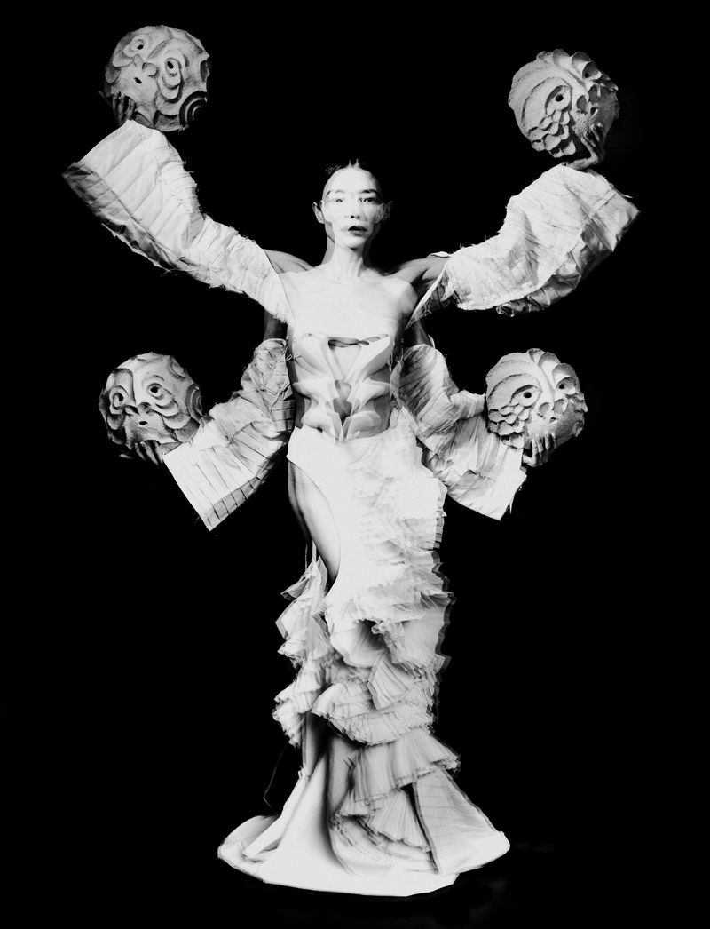 Model wearing a sculptural white ruffled dress with multi-arm extensions holding textured masks against a solid black background.