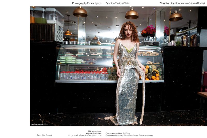 Model with red curly hair posing in a full-length silver sequined evening gown in front of a food display counter inside a cafe.