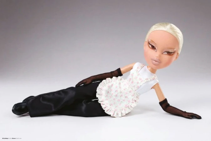 Bratz doll with sleek blonde hair reclining in black trousers, a floral pinafore apron, and sheer black gloves against a grey background.